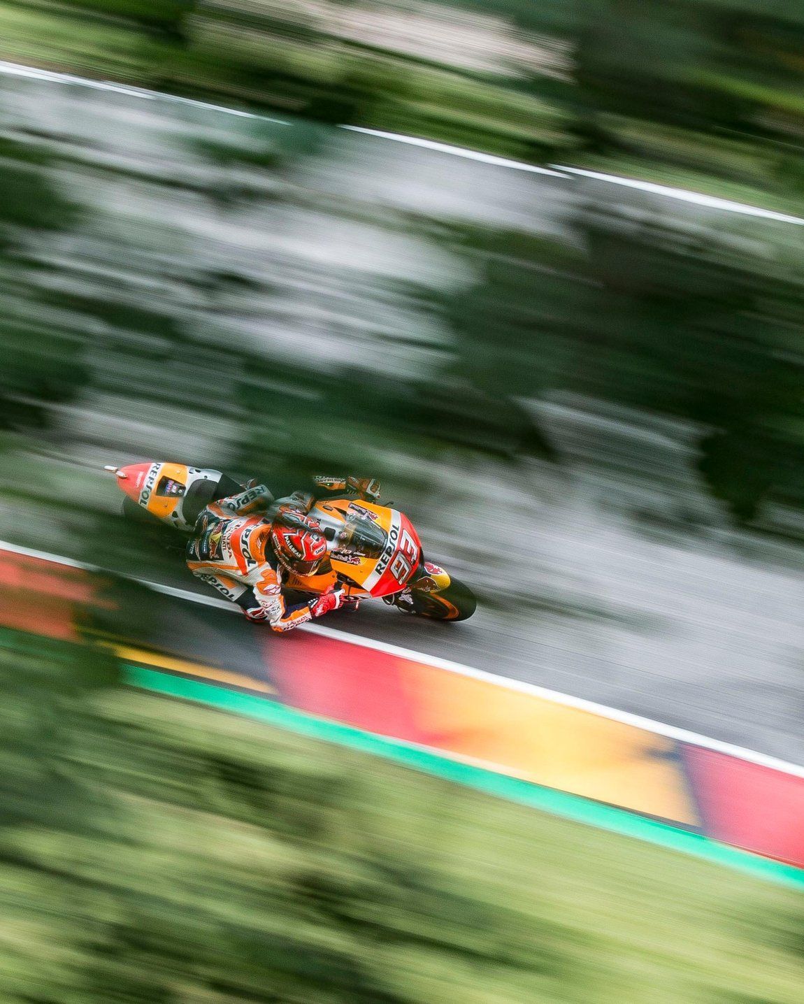 Motorcoureur racet over een natte baan. De wazige beelden tonen de snelheid; de motor heeft een kleurrijk oranje en rood design.
