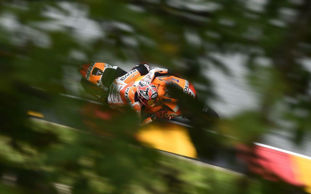 Motorrace op een circuit, gedeeltelijk verhuld door wazig groen gebladerte. Oranje, zwarte en witte motorfiets.