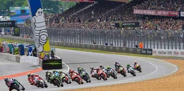 Motorrace op een bochtige baan, waarbij veel motoren langs een Michelin-bord racen en toeschouwers op de achtergrond.