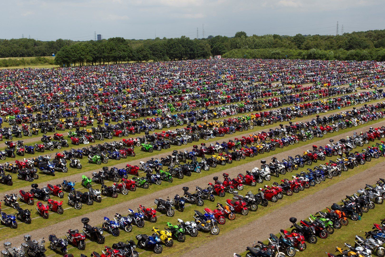 Grote bijeenkomst in de buitenlucht met honderden motoren, geparkeerd in rijen op het gras. Groene bomen en een blauwe lucht op de achtergrond.