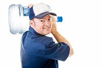Man carrying a large water jug on his shoulder, wearing a blue shirt and cap.