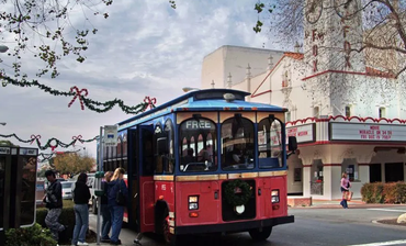 A red and blue trolley loading passengers in front of a theater decorated for the holidays.