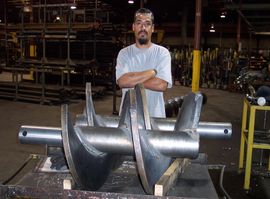 Man in a factory with arms crossed, standing behind a large metal industrial component on a table.