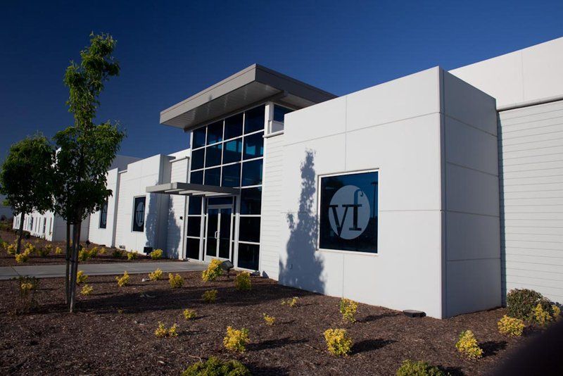 White building with a dark-framed entrance, a logo, and multiple windows; blue sky overhead, landscaping in the foreground.