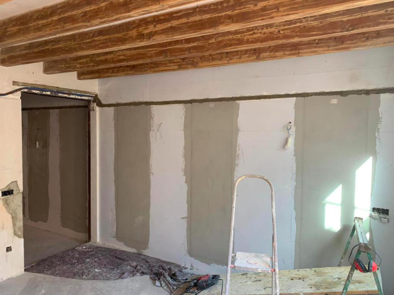 Interior de una habitación en renovación; paneles de yeso y vigas visibles, con una escalera y herramientas presentes.