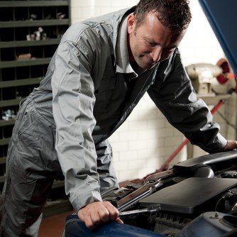 Mechanic in gray jumpsuit working on car engine, using a wrench in a garage.