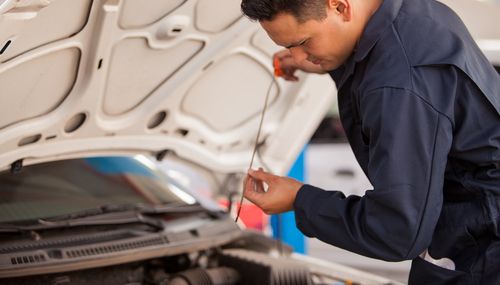 Mechanic in blue jumpsuit checks car's oil dipstick under the hood.