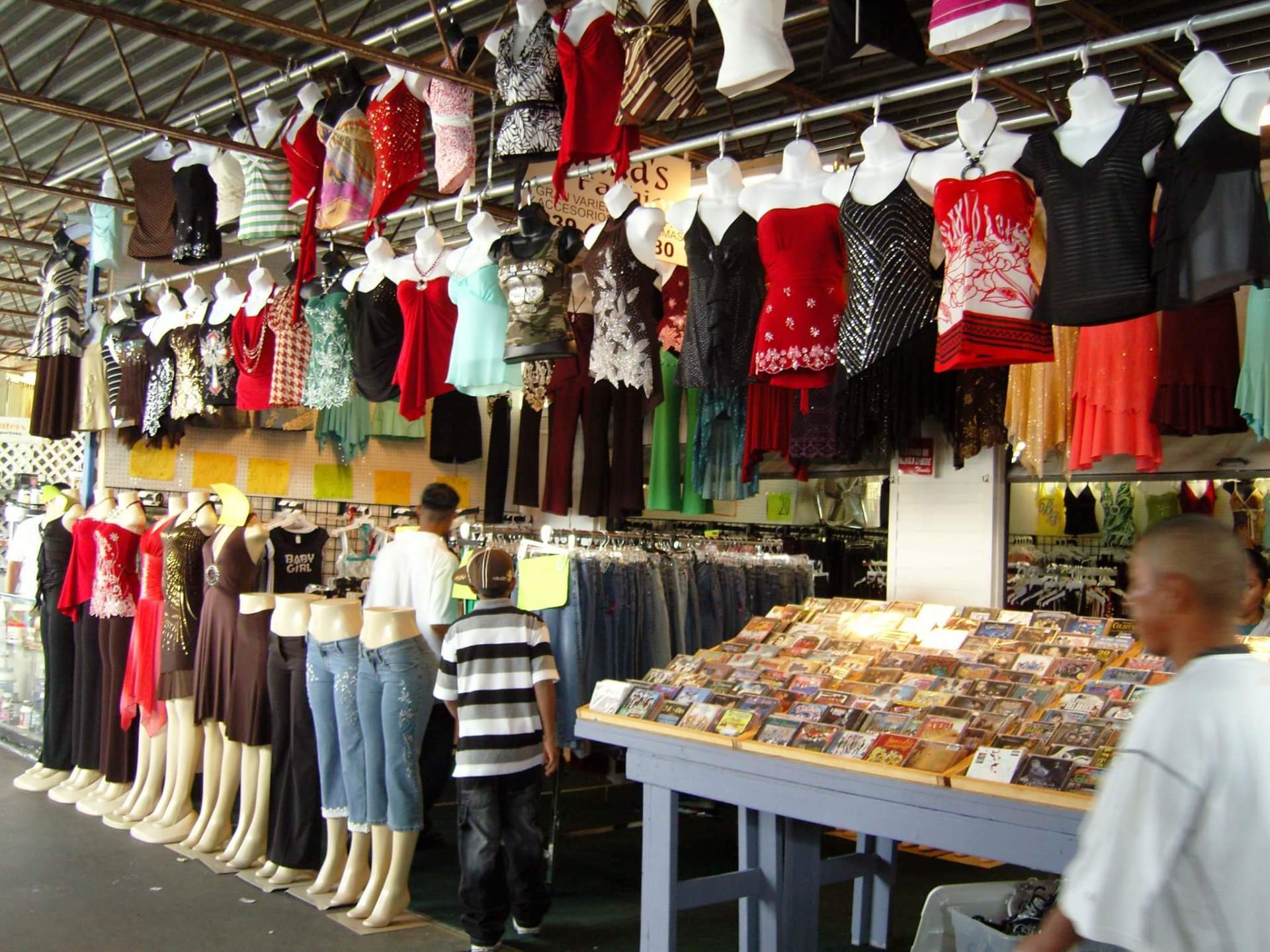 Clothing market stall with mannequins displaying clothes; customers browse items.