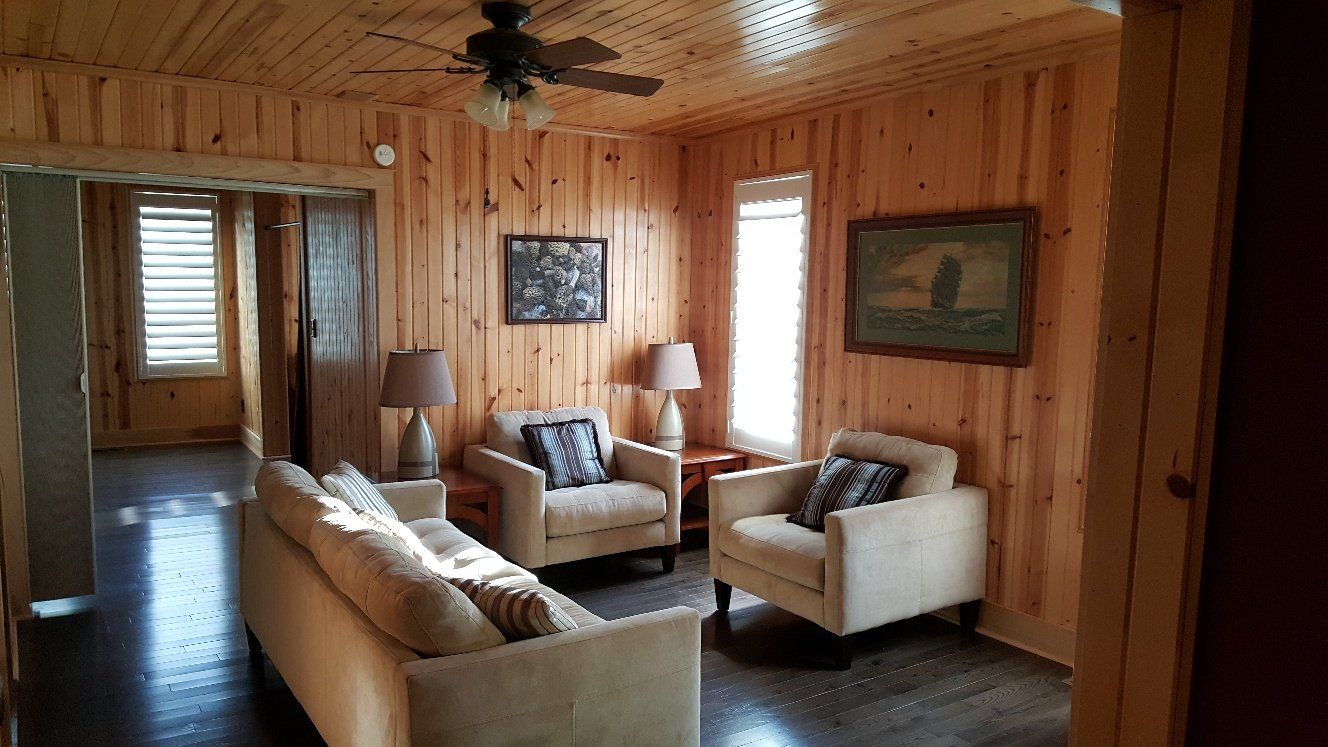 Living room with wood paneled walls, a couch, two armchairs, and a ceiling fan.