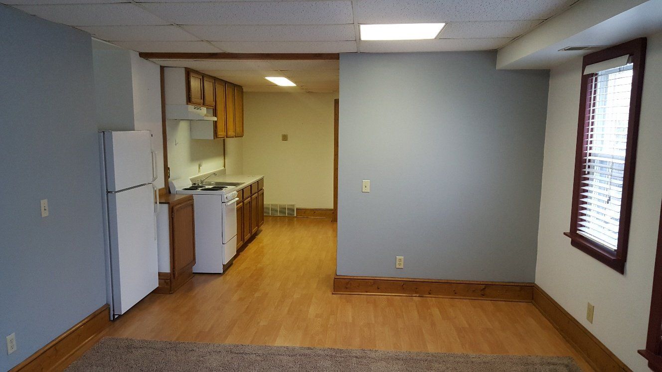 Interior of an apartment with a kitchen, refrigerator, and window. Light blue walls and wood floors.