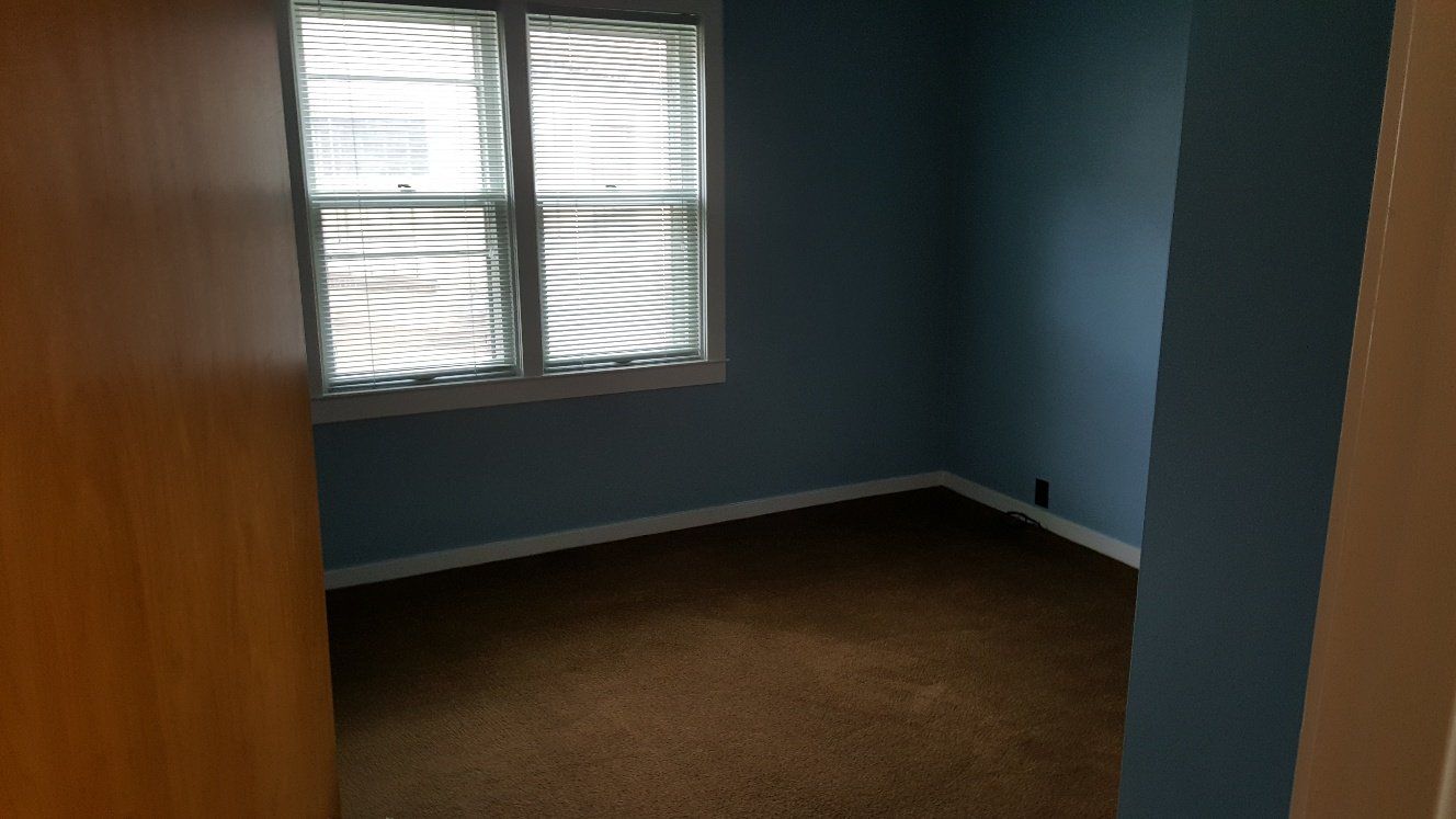 Empty blue-walled room with brown carpet, a window with blinds, viewed from a doorway.