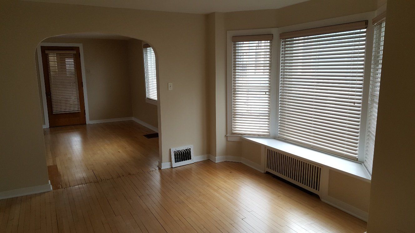Empty living room with wood floors, bay window with blinds, archway to another room.