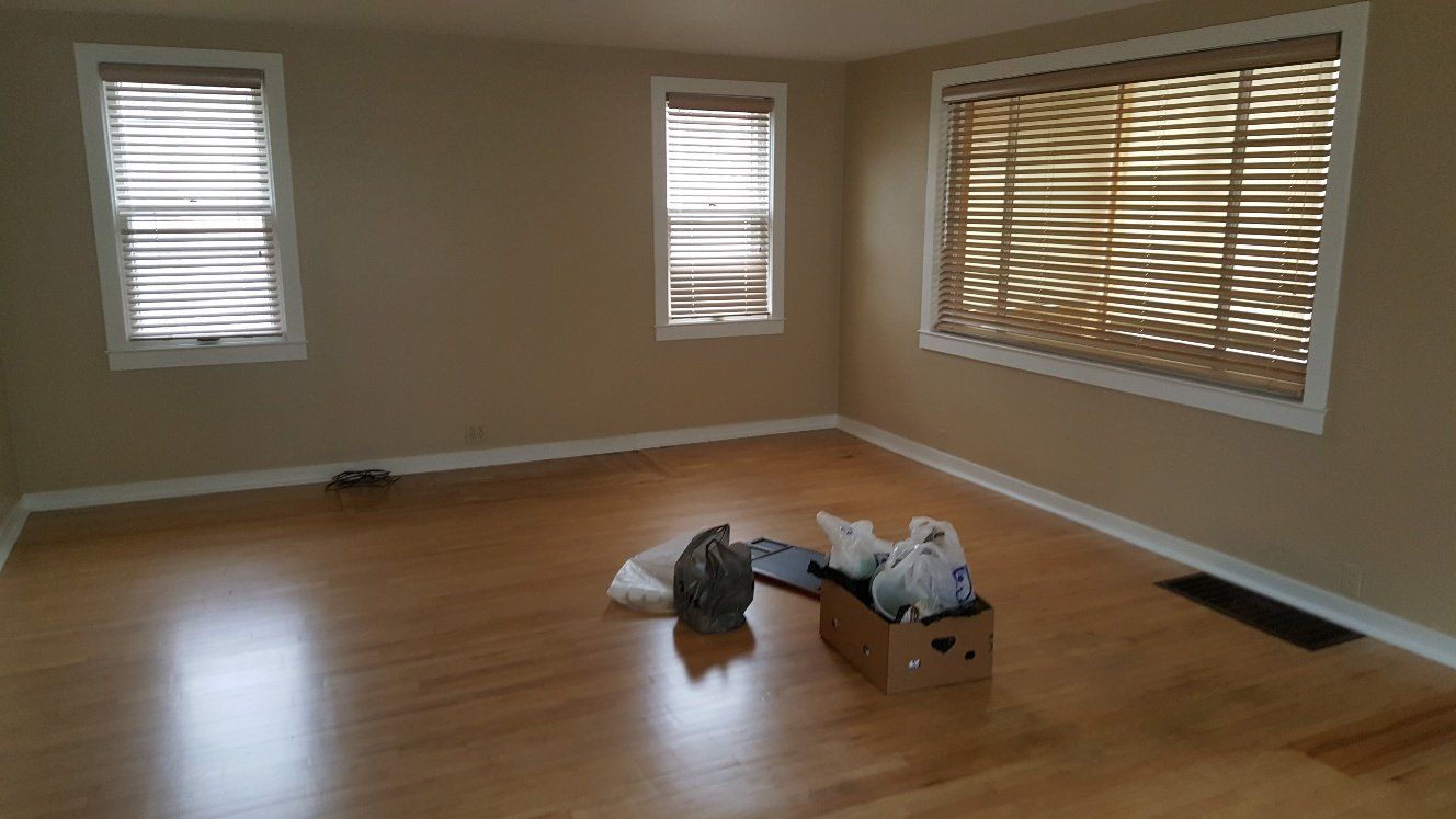 Empty room with hardwood floors, beige walls, and three windows with blinds. Box and two cats on floor.