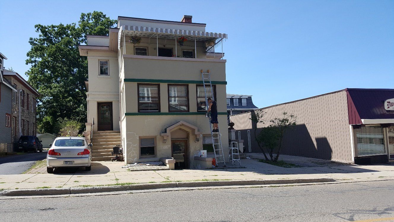 Three-story beige building with green trim, person on a ladder, a car parked in front, clear blue sky.