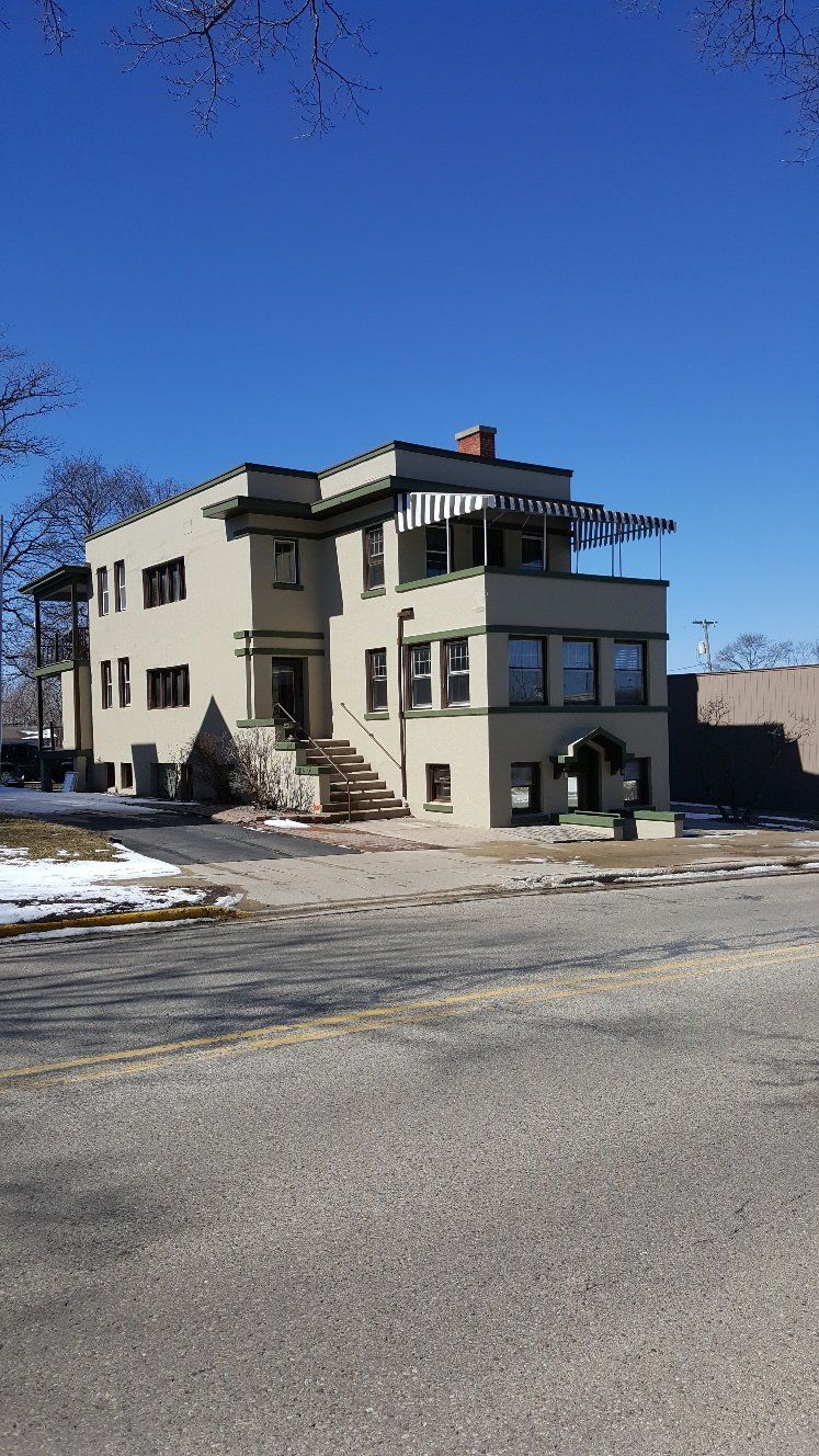 Three-story tan building with a flat roof and a small porch on a sunny day. Snow patches visible.