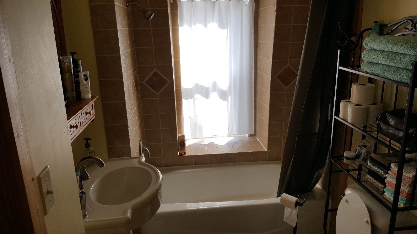 Bathroom with a window, sink, and bathtub. Beige tiles and walls, with a black shower curtain and a metal shelf.