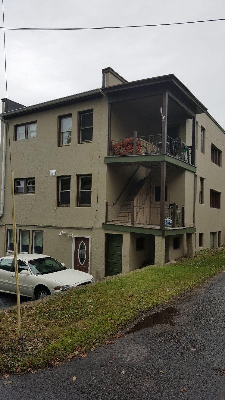 Tan apartment building with balconies, stairs, and a parked white car on a sloped street.