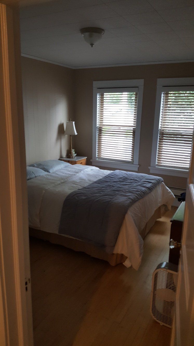 Bedroom with bed, windows, lamp, and fan. Wooden floor and beige walls.