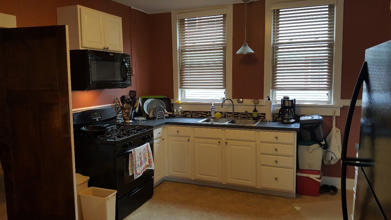 Kitchen with black appliances, white cabinets, and two windows with blinds. Reddish-brown walls.