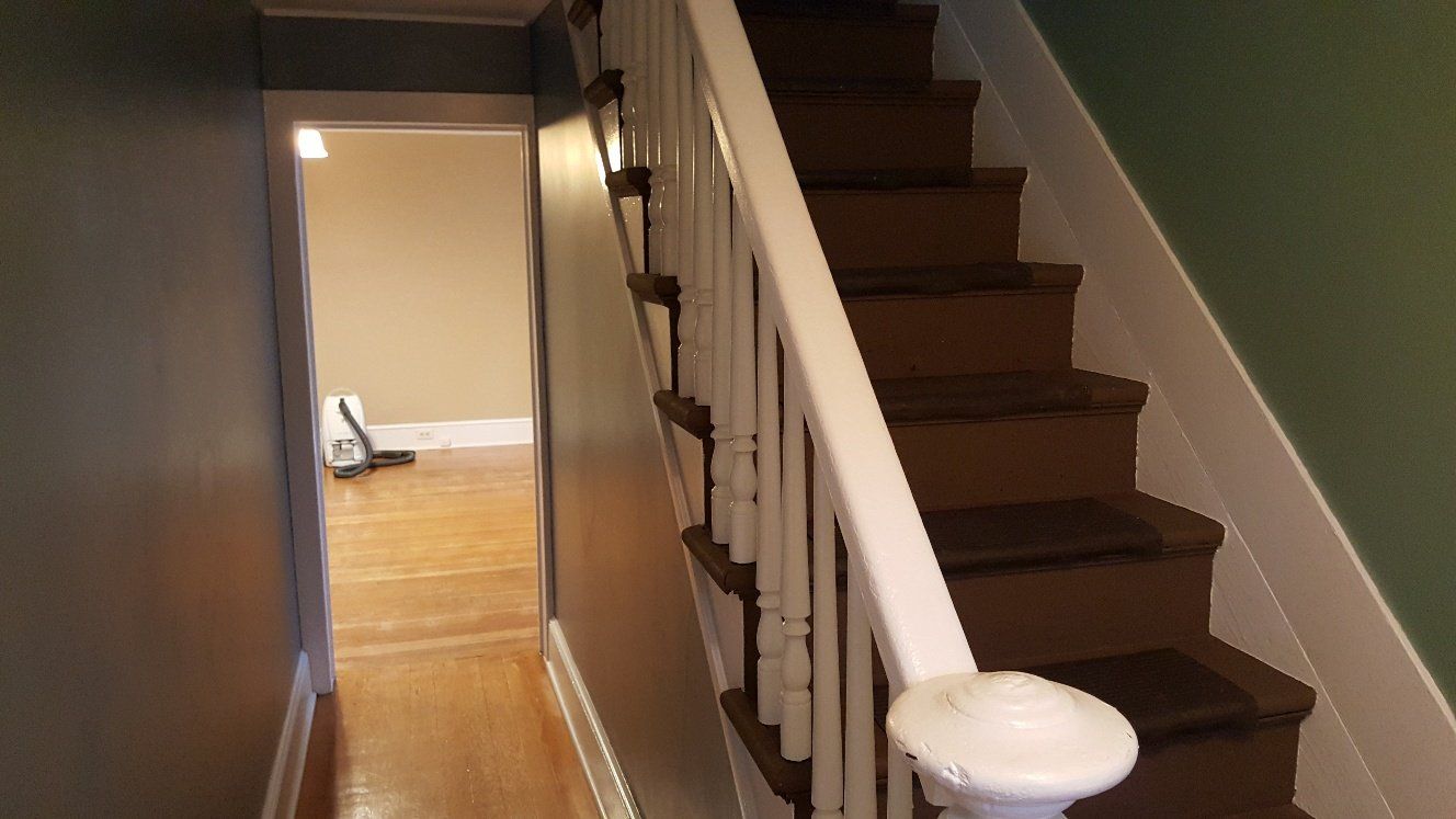 Staircase with brown steps and white railing, leading to a hallway with wooden floors and a room with beige walls.