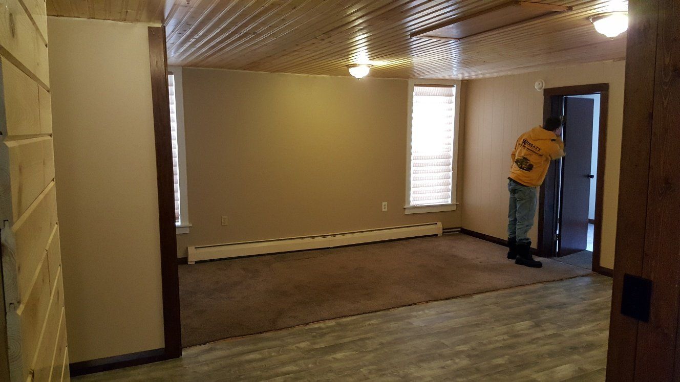 Room interior with person standing in doorway, brown walls, carpet, and exposed wood ceiling.