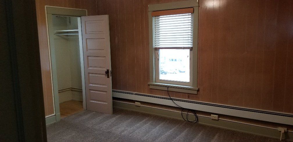 An empty room with a closet, window, and baseboard heating. The walls are paneled and painted reddish-brown.