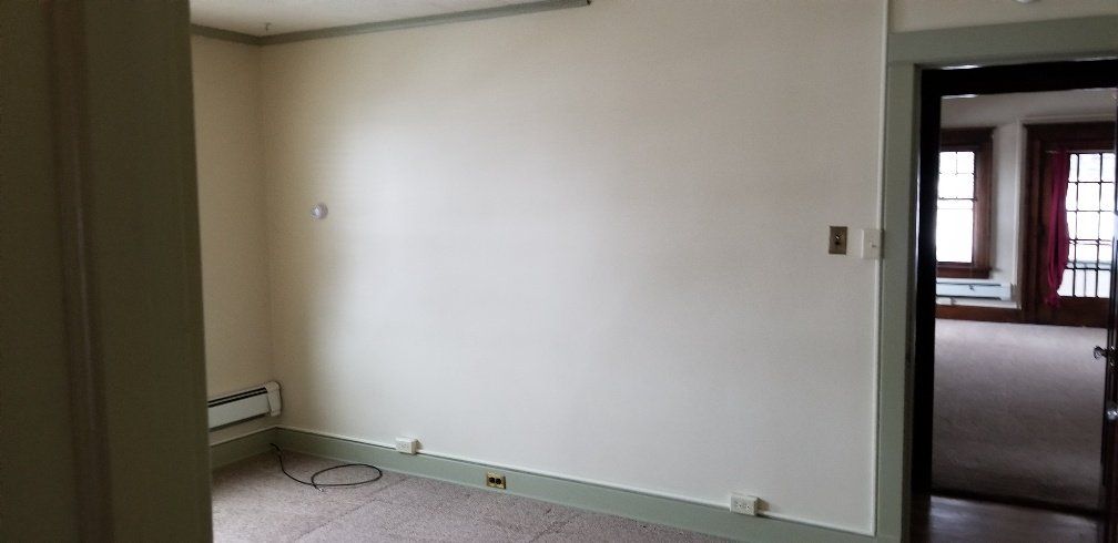 An empty room with light walls and a doorway to another room. Green trim and beige carpet.