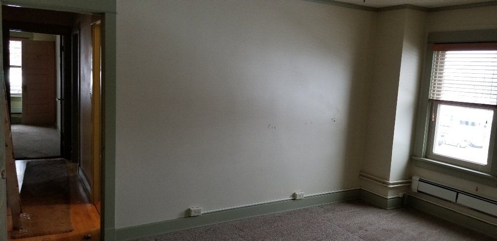 Empty room with beige walls, a window with blinds, and a doorway to another room.
