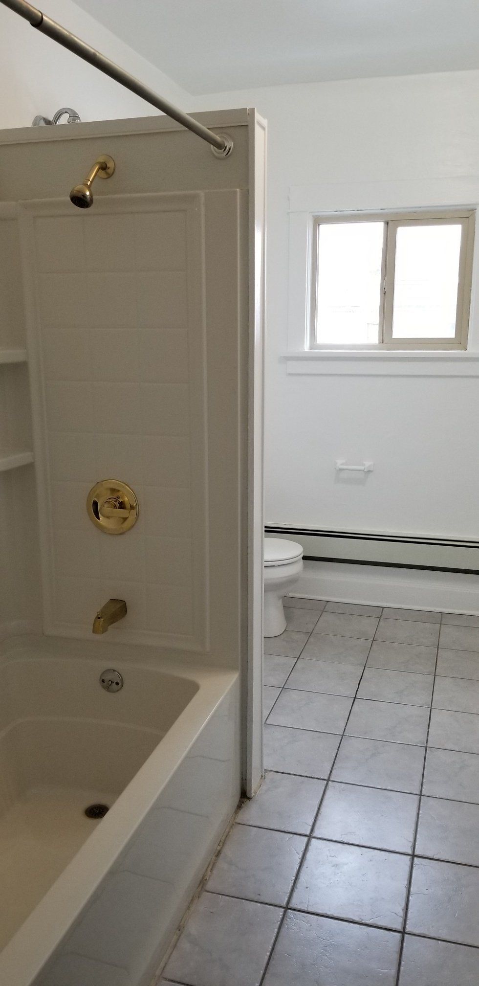 Bathroom with white tub, shower fixture, toilet, window, and gray tiled floor.