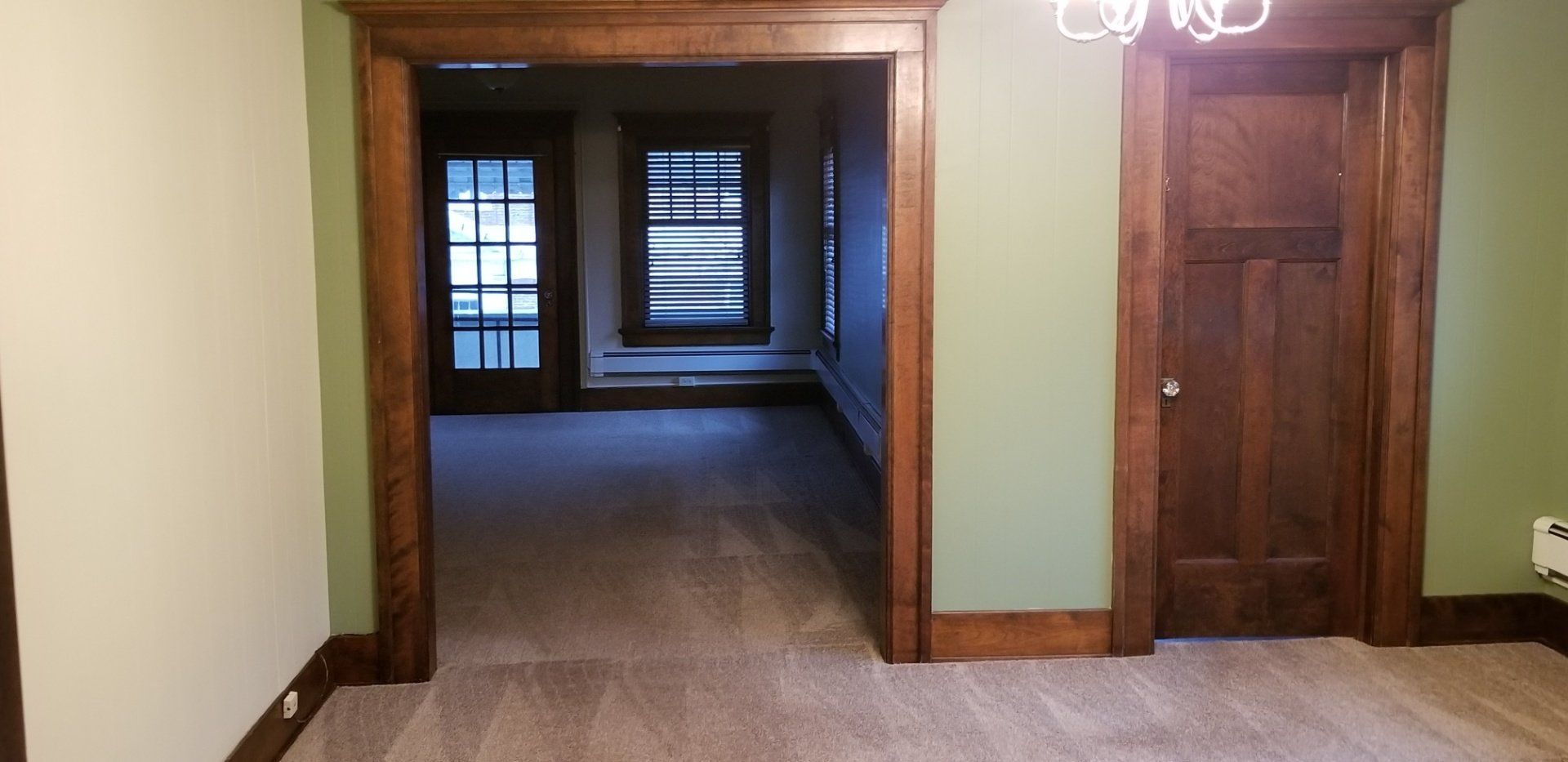 Interior view of a room with green walls, wood trim, doorway to a room with windows and a dark wood door.