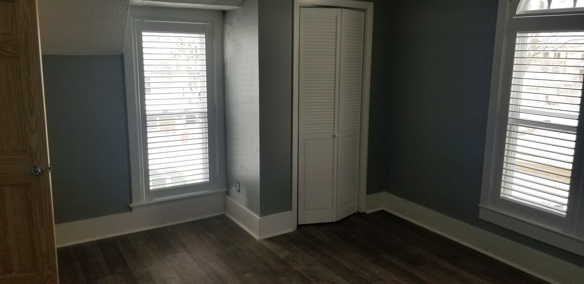 An empty bedroom with gray walls, white trim, and a closed white closet. Two windows with blinds are visible.