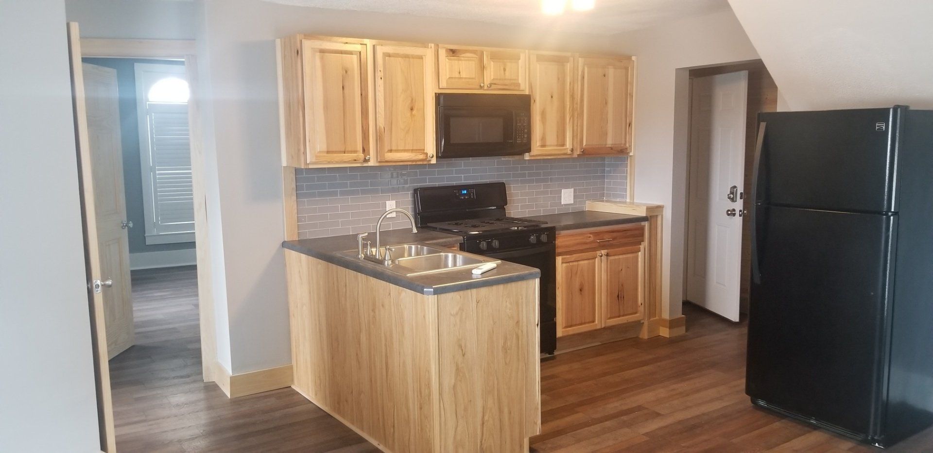 Kitchen with light-colored cabinets, black appliances, and gray countertops. Wooden floors and walls are visible.