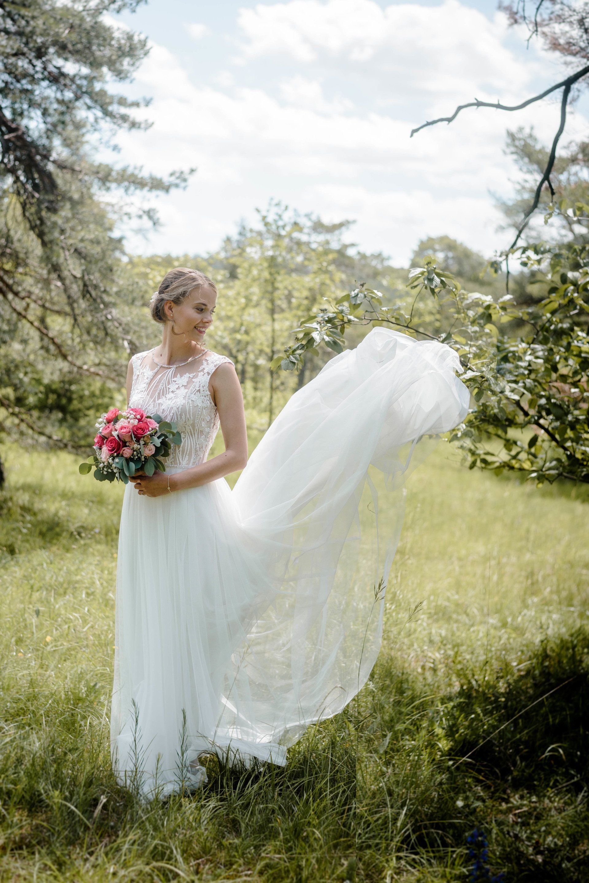 Eine Braut im Brautkleid steht mit einem Blumenstrauß in der Hand auf einem Feld.
