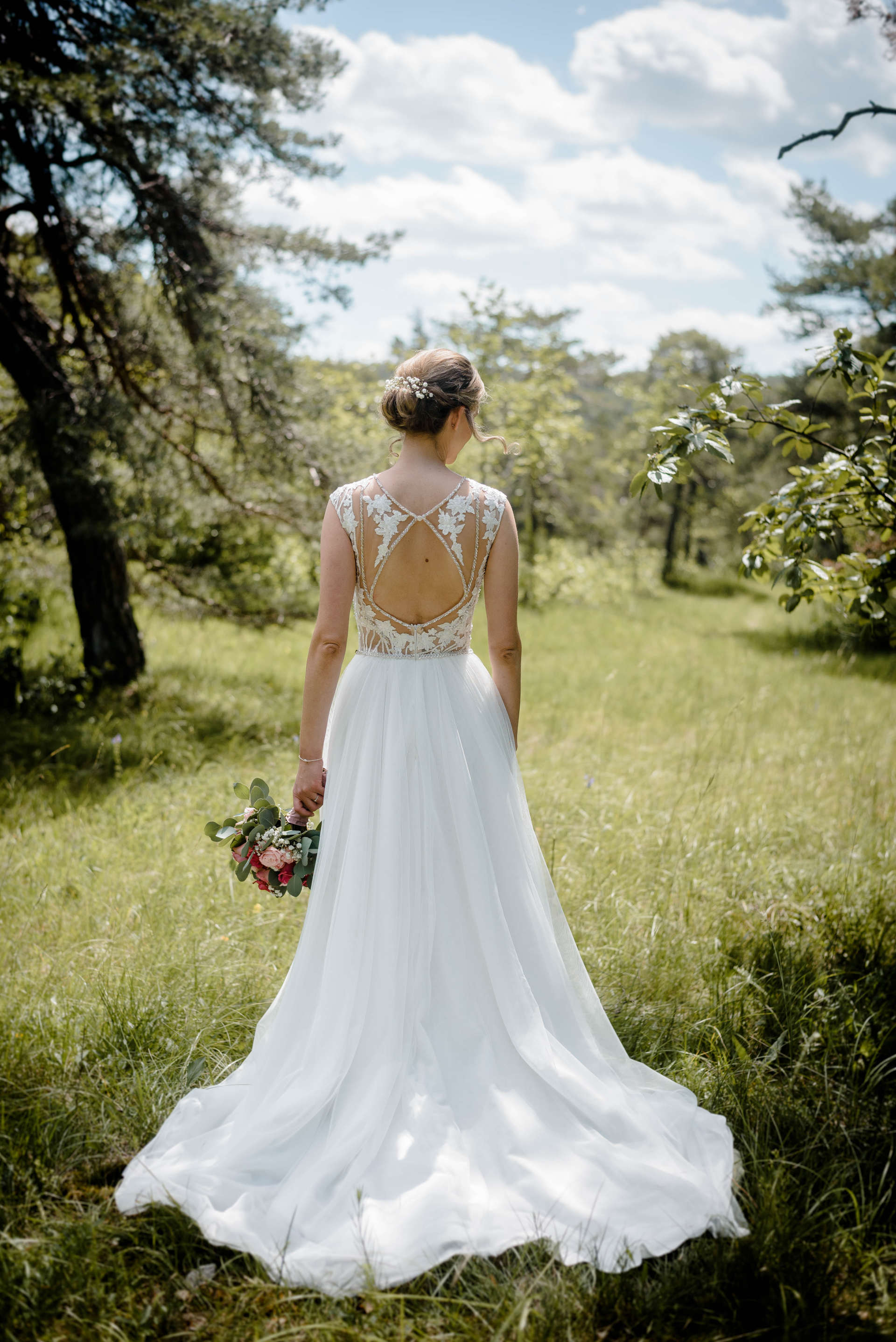 Eine Braut im Brautkleid steht mit einem Blumenstrauß in der Hand auf einem Feld.