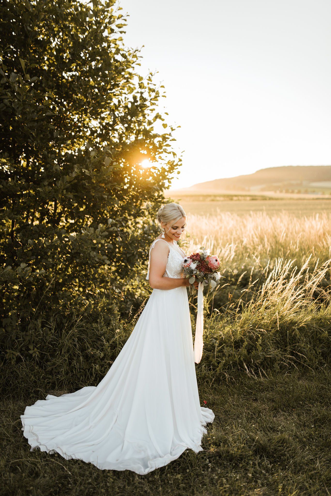 Eine Braut im Brautkleid steht mit einem Blumenstrauß in der Hand auf einem Feld.
