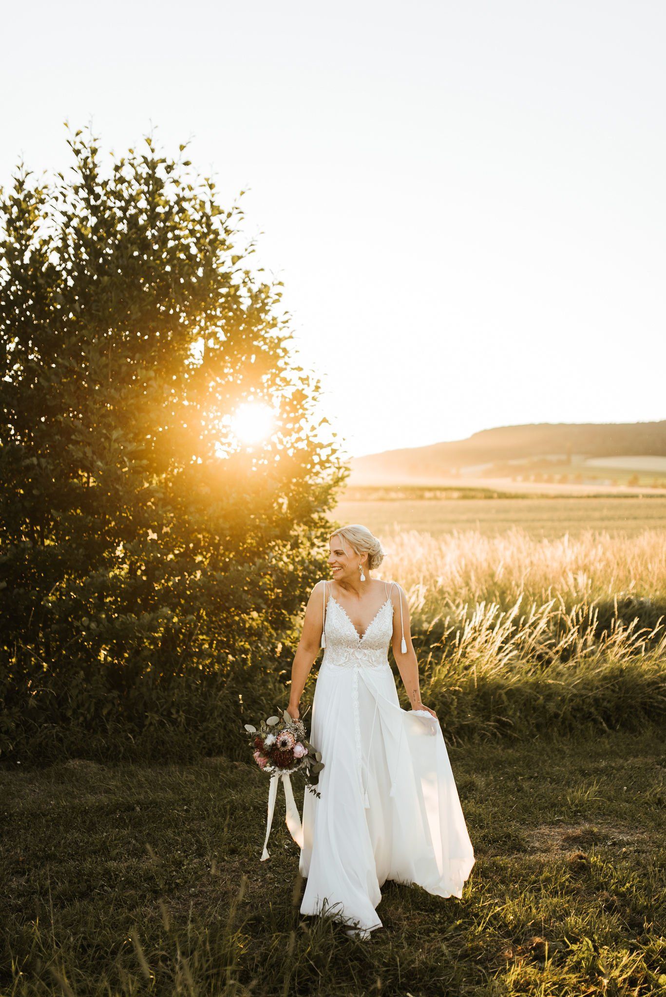 Eine Frau in einem Brautkleid steht mit einem Blumenstrauß in der Hand auf einem Feld.