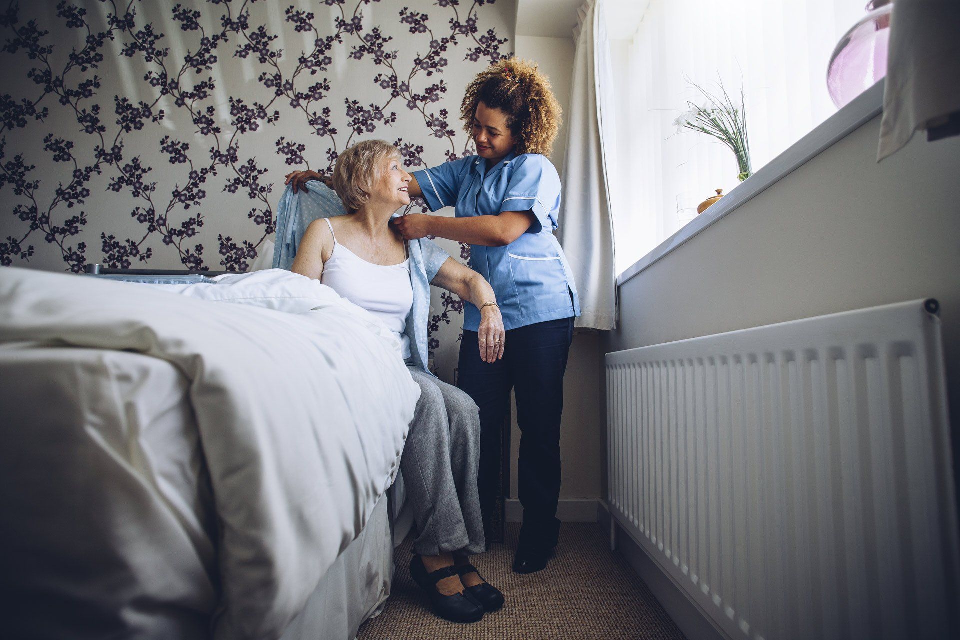 home-caregiver-helping-a-senior-woman-get-dressed-in-her-bedroom-482413720-1920w
