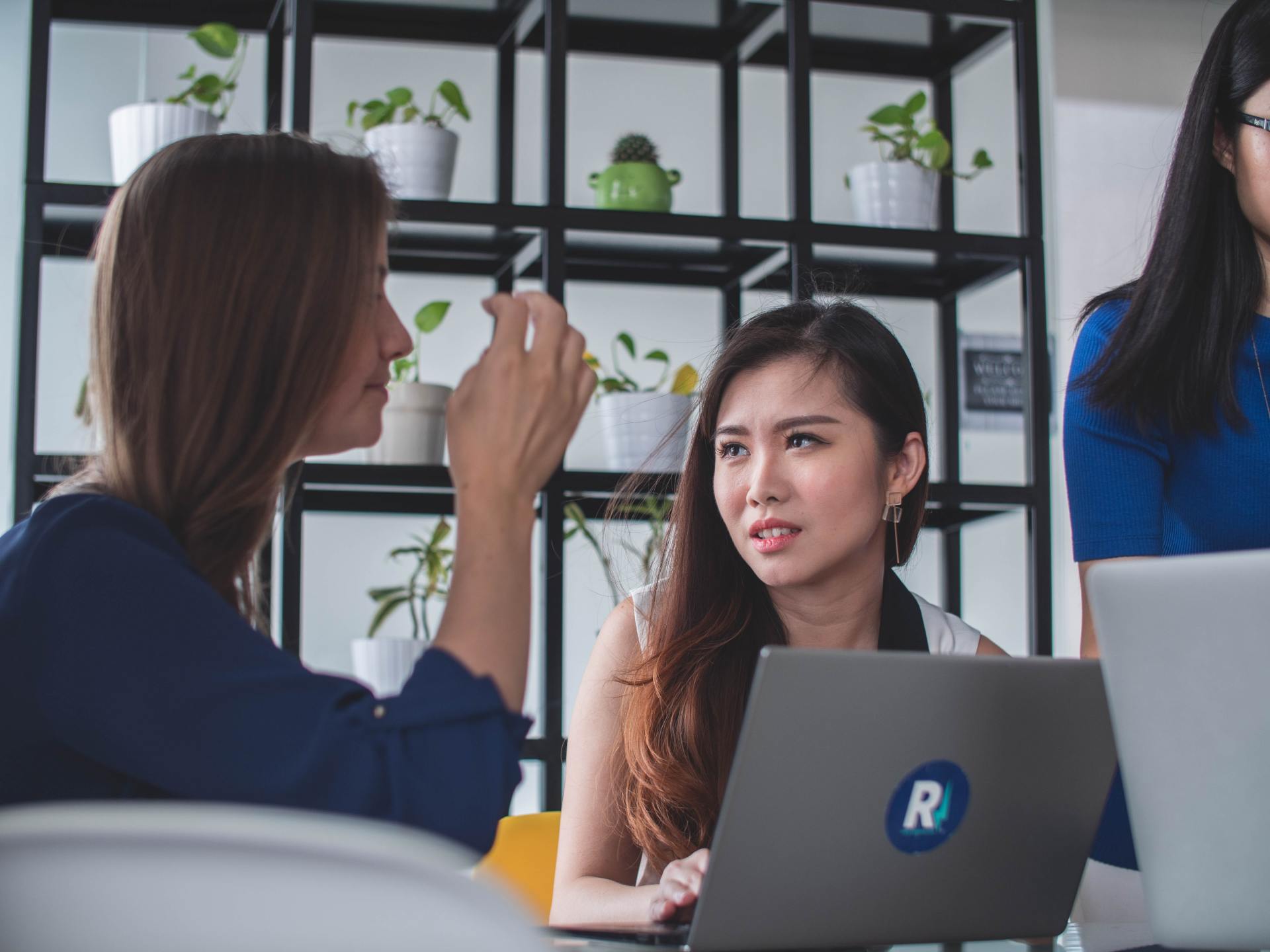 Three women are sitting at a table with laptops and talking to each other.
