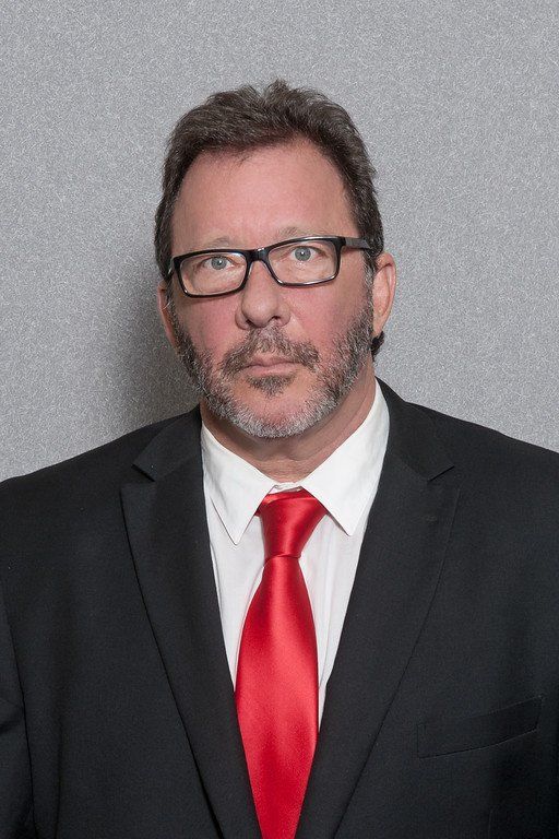 Man in a black suit and red tie, wearing glasses, posed against a gray backdrop