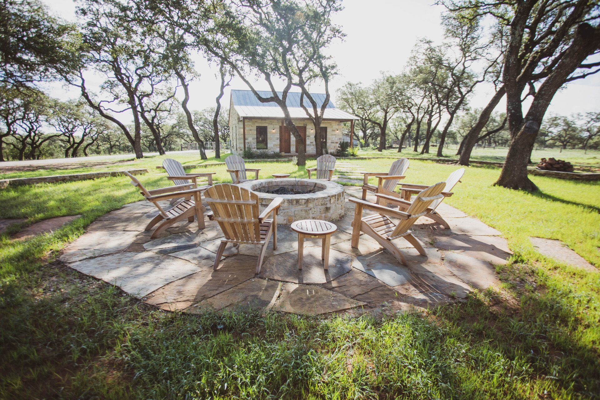 Fire pit with wooden chairs on a stone patio in a grassy area; small stone building in the background.