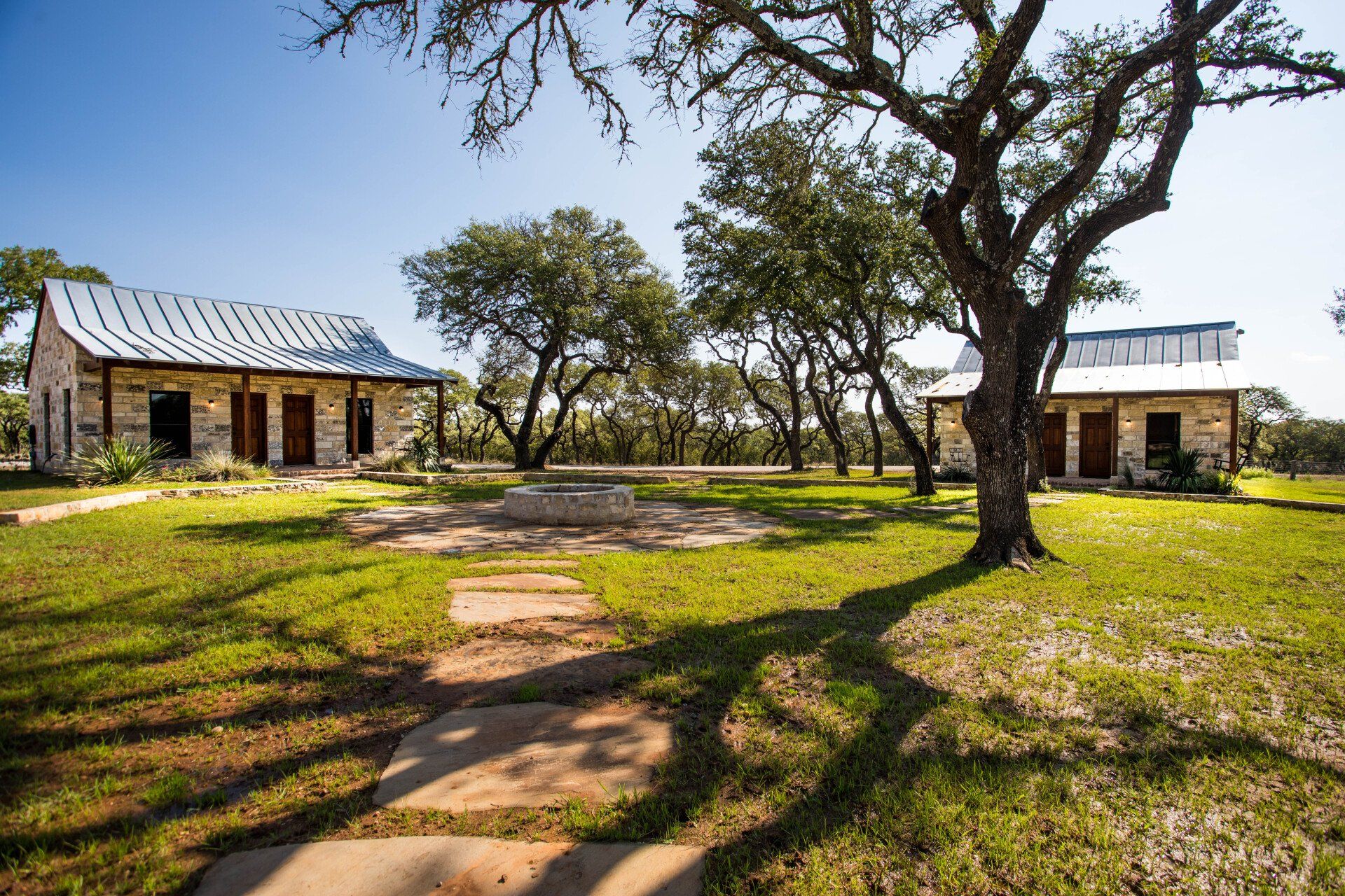 Two stone cabins with metal roofs on a grassy lawn, trees casting shadows.