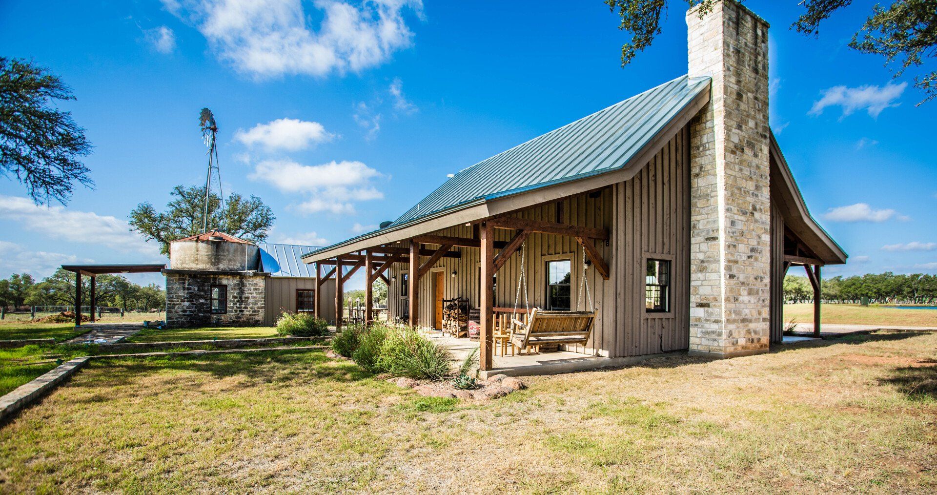 Rustic wooden cabin with a stone chimney, porch, and blue metal roof on a sunny, grassy field.