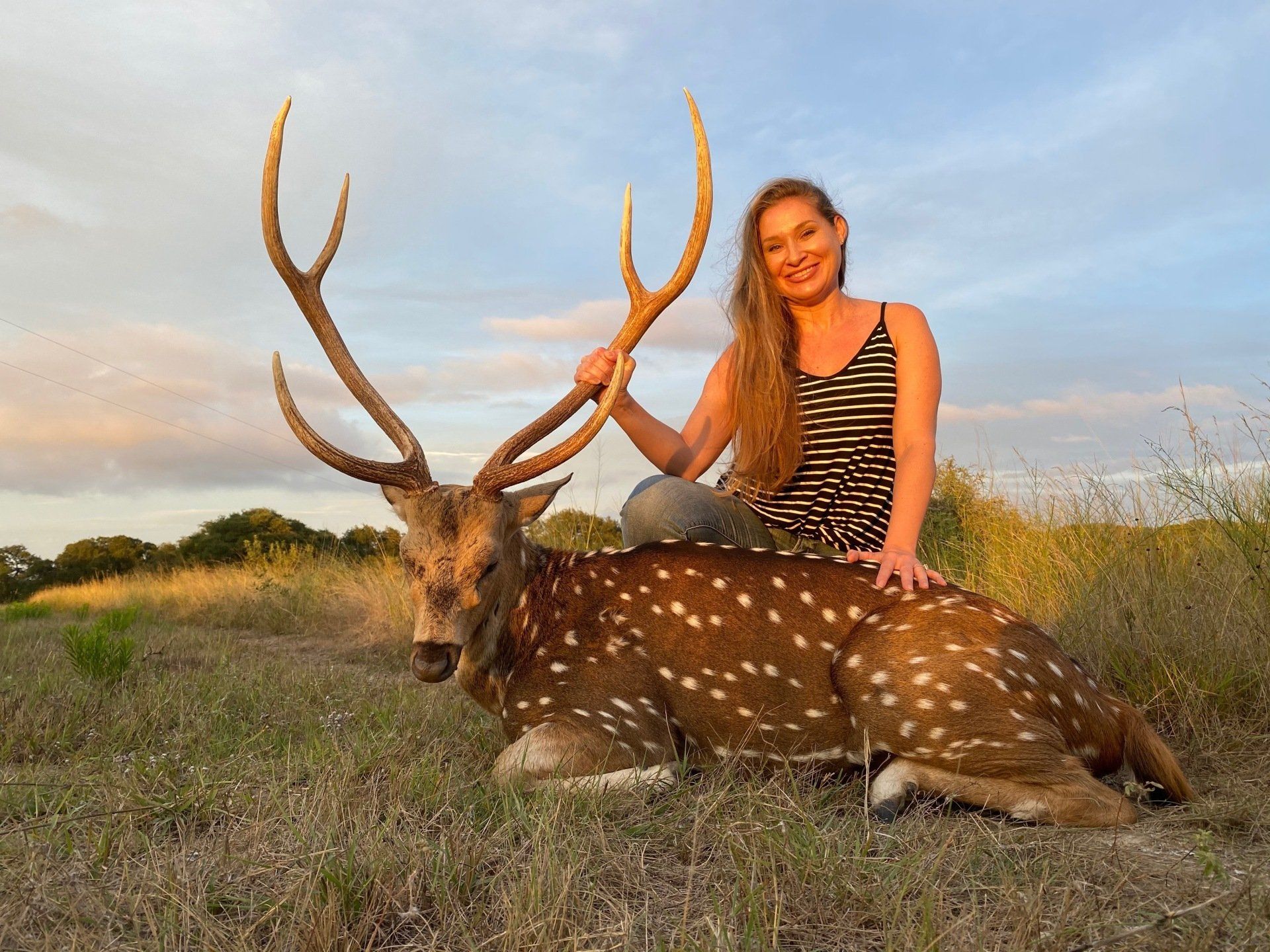 Woman posing with a spotted deer she hunted, outdoors. She is smiling and touching the antlers.