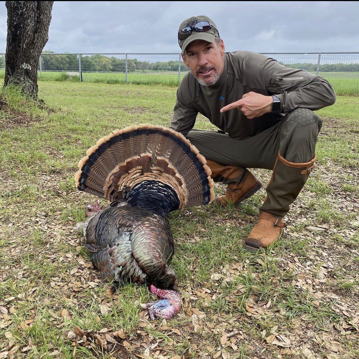Hunter kneeling next to a harvested turkey, pointing. He's in field, wearing green and brown clothing.
