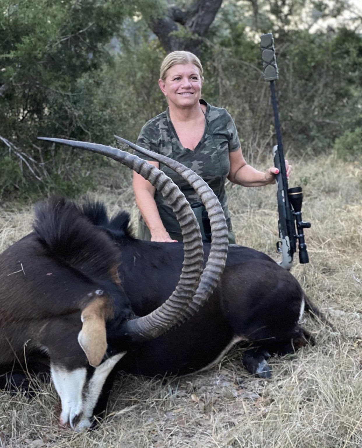 Woman in camo smiles, poses with a dead sable antelope and rifle in a grassy field.