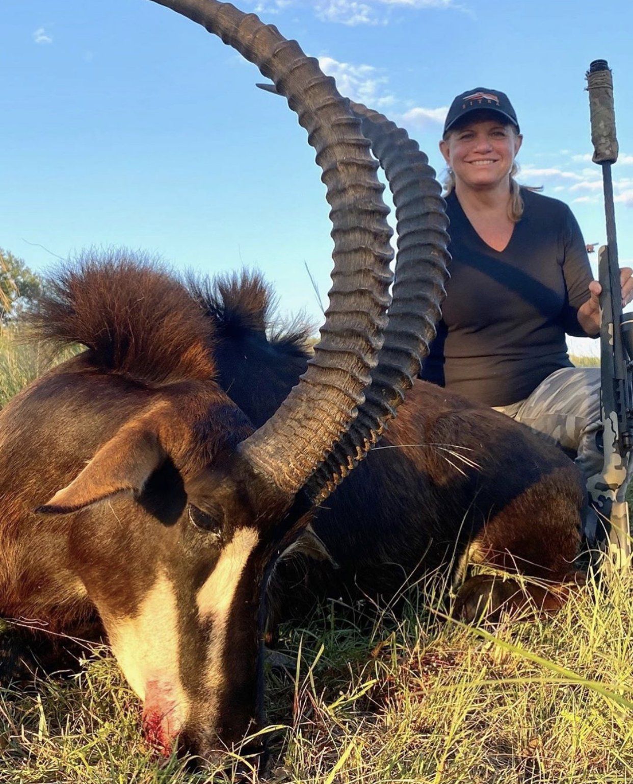 Woman kneels next to a dead sable antelope, holding a rifle. Outdoors, tall horns, dark fur.