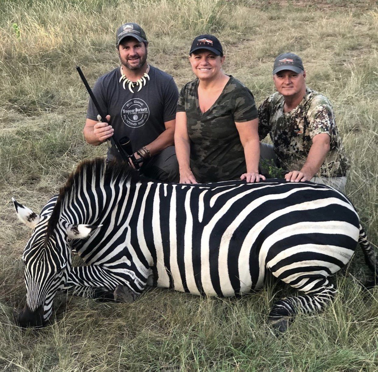 Three people pose with a dead zebra in grassy field. Man holds rifle. All smile.