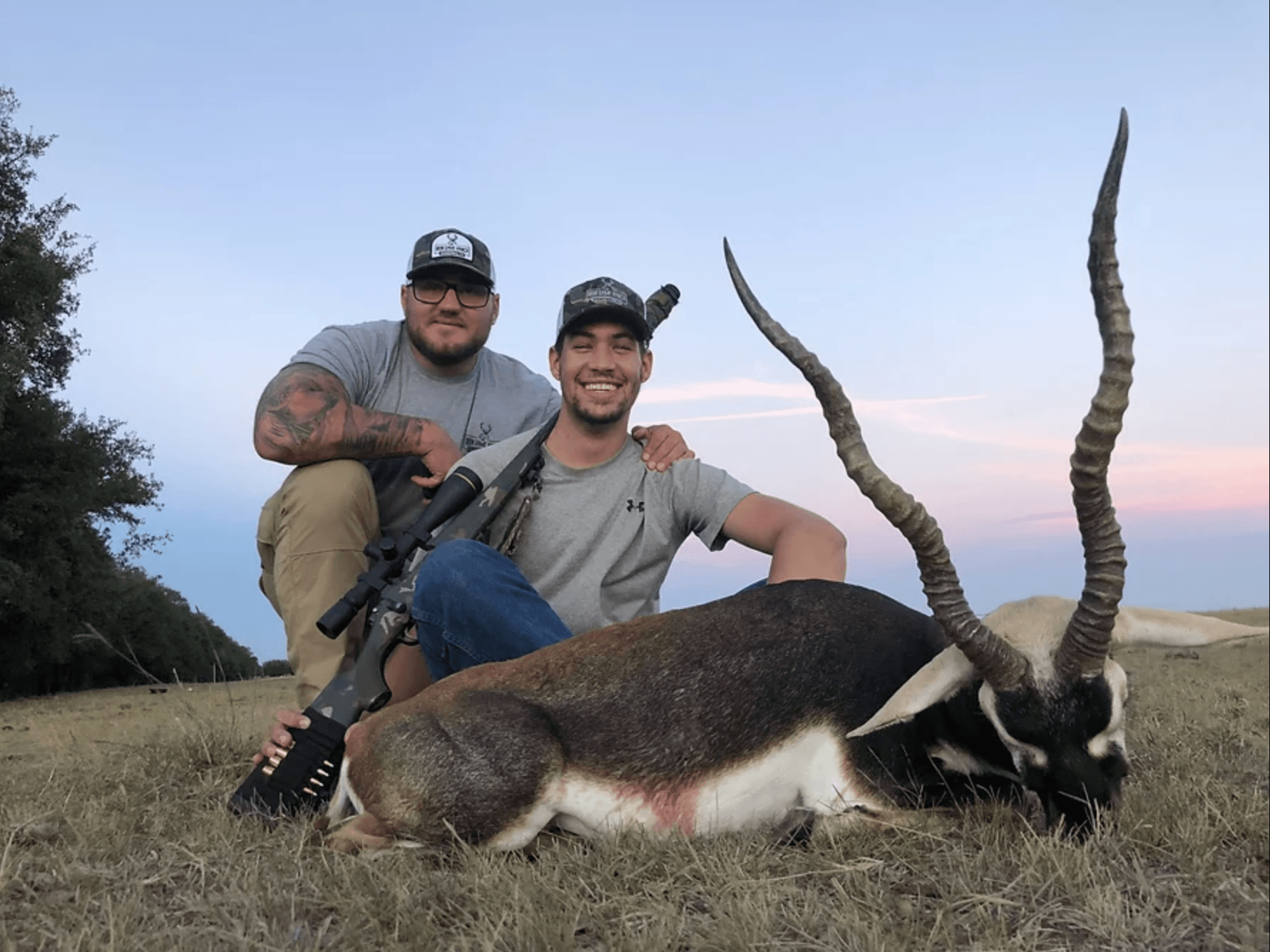 Two men pose with a dead blackbuck antelope in a field. One man holds a rifle.