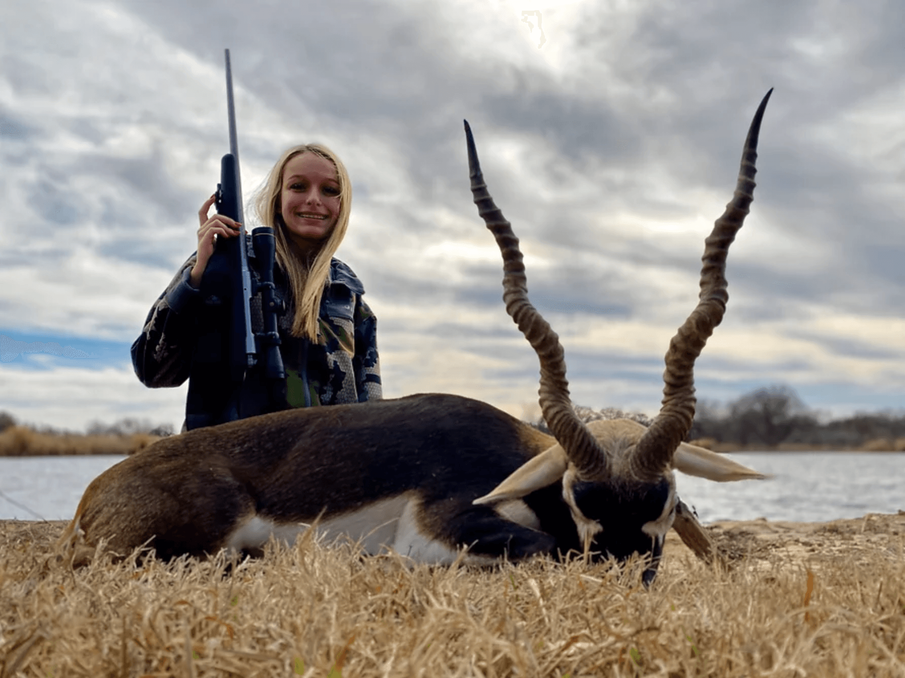 Woman in camo with a rifle stands next to a blackbuck on a grassy bank by water.