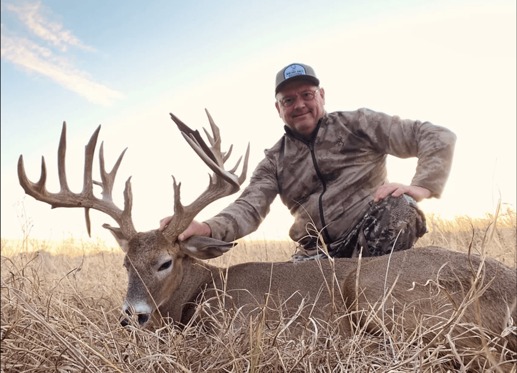 Hunter kneels beside a large buck with impressive antlers in a field. He smiles, petting the deer.